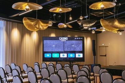 Salle de casino avec table de jeu vide, chaises alignées, machines à sous illuminées et éclairage suspendu moderne.