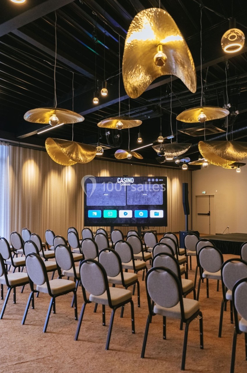Salle de conférence avec rangées de chaises grises, écran de projection allumé et luminaires dorés suspendus.