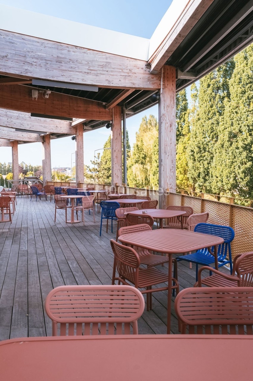 Terrasse en bois avec des tables et chaises colorées, entourée d'arbres et partiellement couverte par une structure en bois.