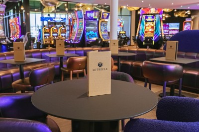 Salle de casino avec table de jeu vide, chaises alignées, machines à sous illuminées et éclairage suspendu moderne.