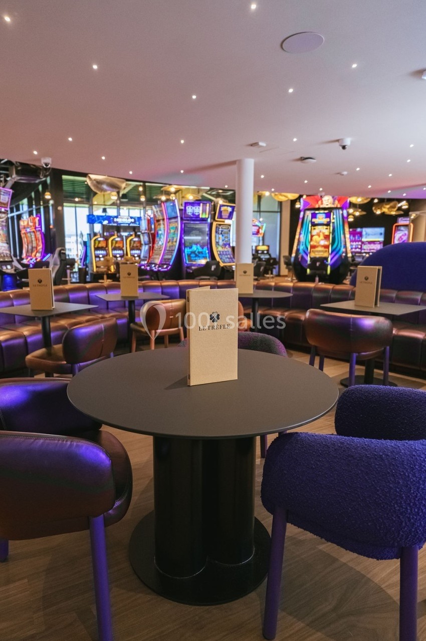 Salle de casino avec machines à sous illuminées en arrière-plan et table ronde entourée de fauteuils au premier plan.