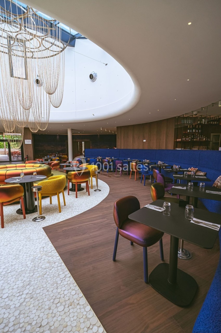 Salle de restaurant moderne avec chaises colorées, tables dressées et grand lustre suspendu sous une verrière.