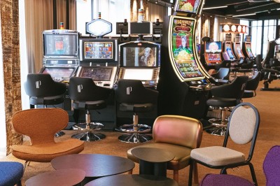Salle de casino avec table de jeu vide, chaises alignées, machines à sous illuminées et éclairage suspendu moderne.
