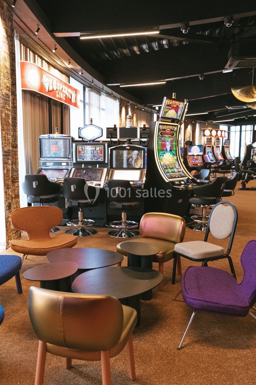 Salle de jeux avec machines à sous alignées et espace détente aménagé avec des fauteuils colorés et des tables basses.