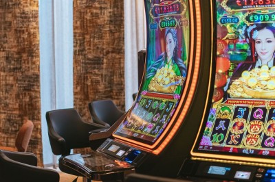 Salle de casino avec table de jeu vide, chaises alignées, machines à sous illuminées et éclairage suspendu moderne.