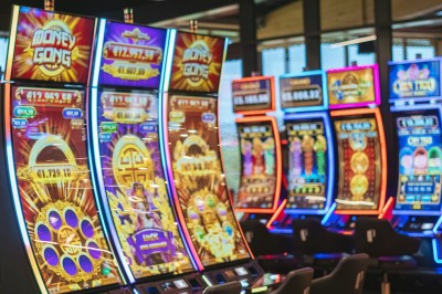 Salle de casino avec table de jeu vide, chaises alignées, machines à sous illuminées et éclairage suspendu moderne.