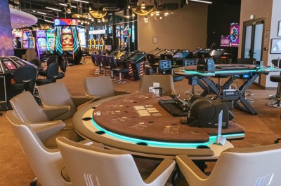 Salle de casino avec table de jeu vide, chaises alignées, machines à sous illuminées et éclairage suspendu moderne.