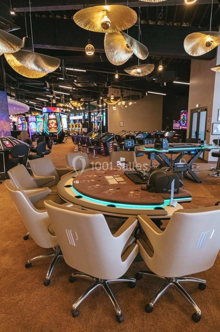 Salle de casino avec table de jeu vide, chaises alignées, machines à sous illuminées et éclairage suspendu moderne.