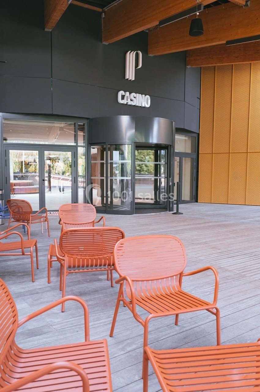 Entrée d'un bâtiment avec une enseigne ’Casino’, terrasse en bois et chaises orange disposées à l'extérieur.