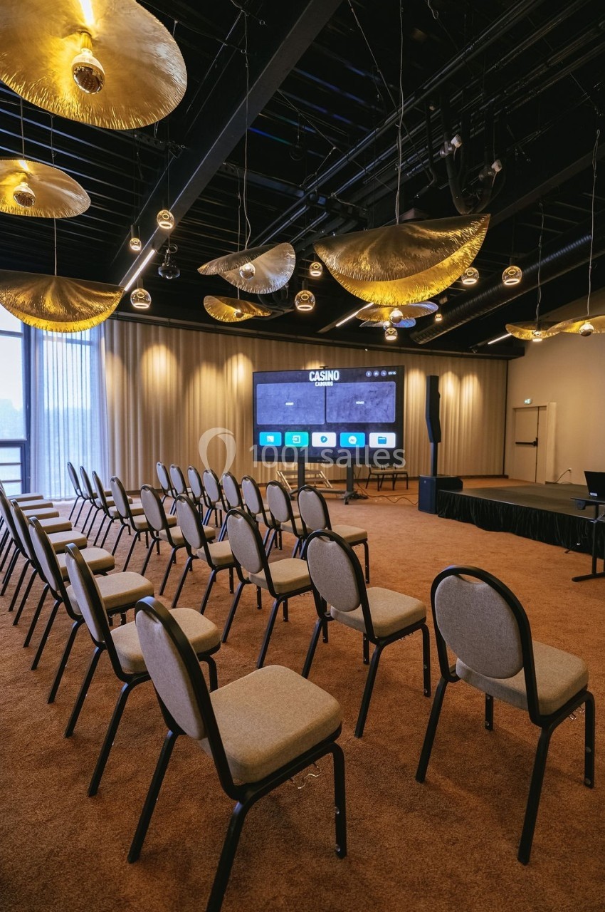 Salle de conférence avec des rangées de chaises vides, un écran de projection allumé et des luminaires suspendus.
