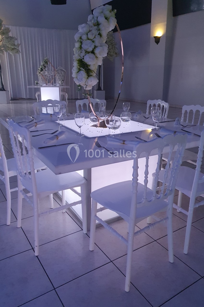 Table élégamment dressée avec des chaises blanches, centre de table floral et éclairage tamisé dans une salle de réception.