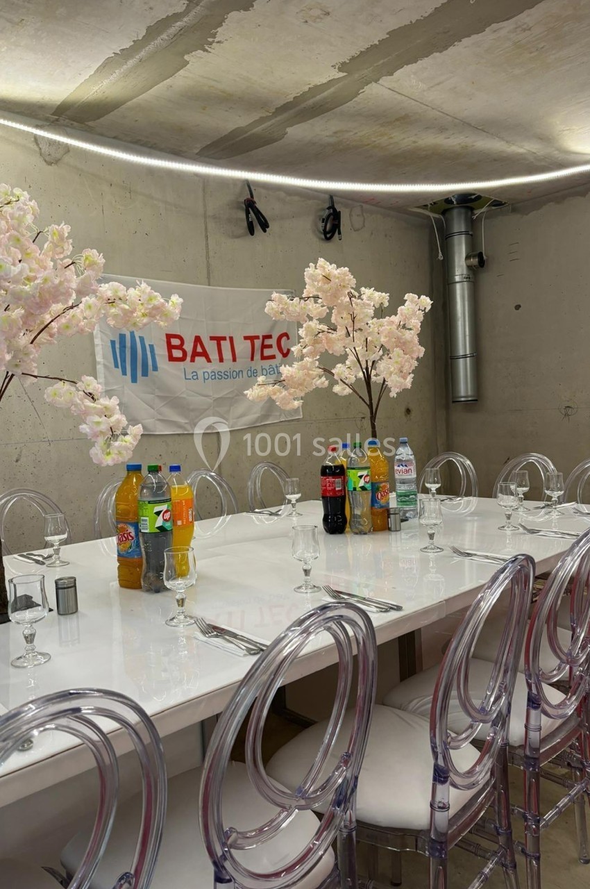 Table dressée avec des chaises transparentes, boissons variées et décorations florales dans une pièce au mur brut.