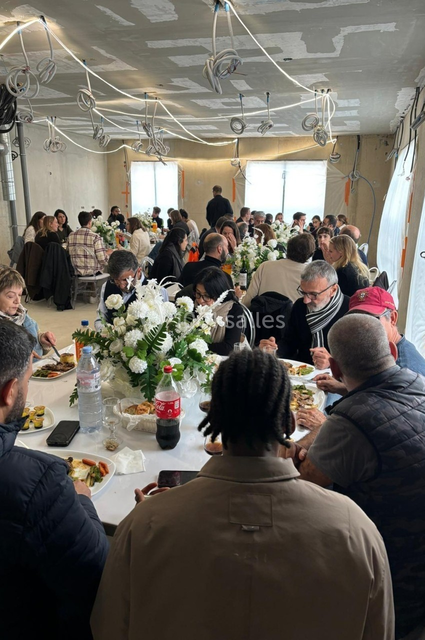 Des convives assis autour de grandes tables décorées de fleurs blanches, partageant un repas dans une salle en travaux.