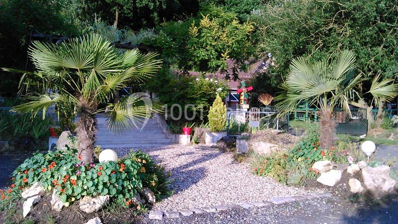 Jardin avec des palmiers, des plantes fleuries, un chemin de gravier et une terrasse entourée de verdure.
