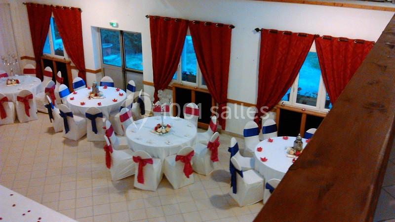 Salle décorée pour un événement, avec des tables rondes, des chaises ornées de nœuds rouges et bleus, et des rideaux rouges.