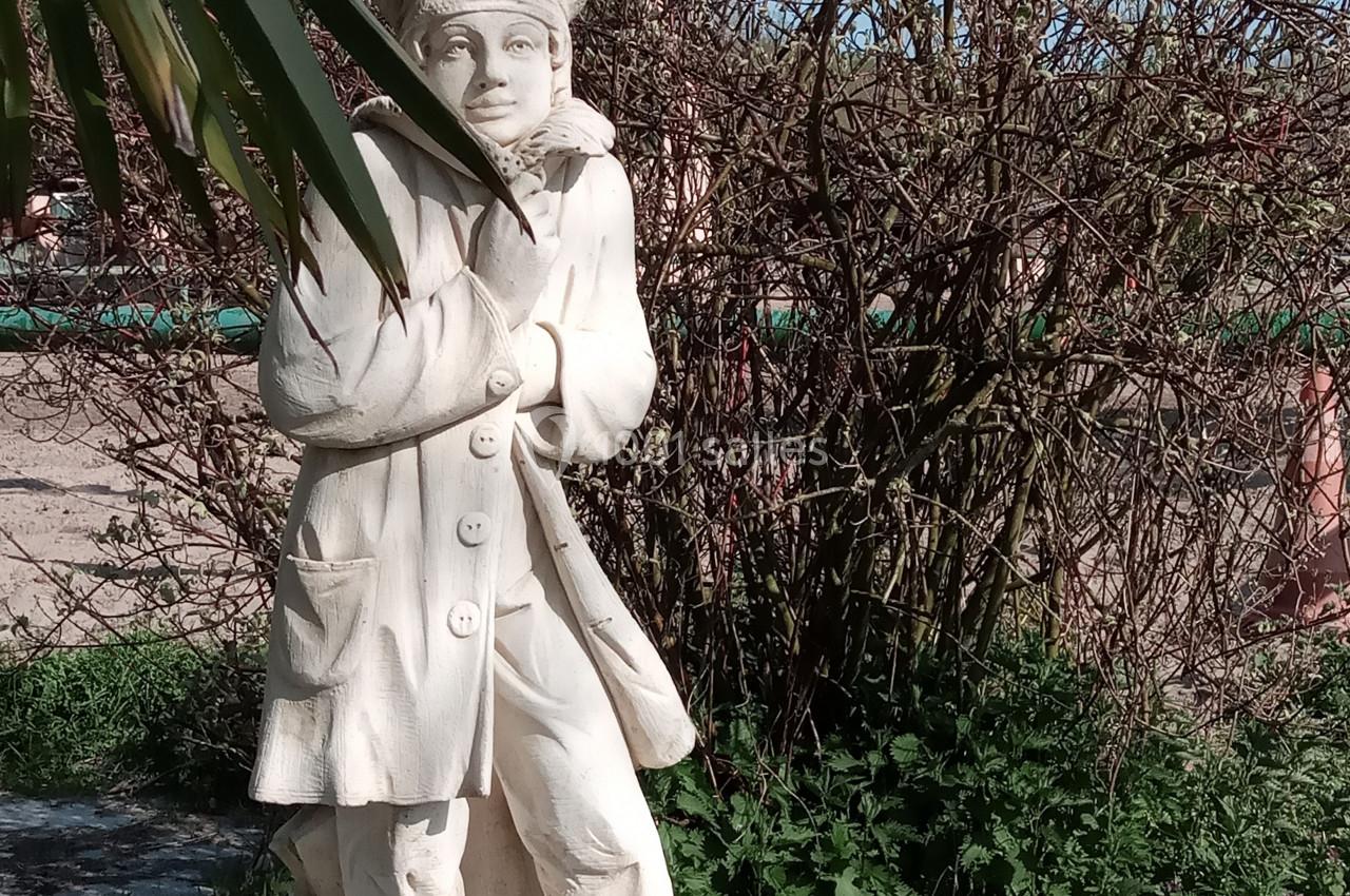 Statue en pierre représentant une personne en manteau et chapeau, entourée de végétation et éclairée par le soleil.