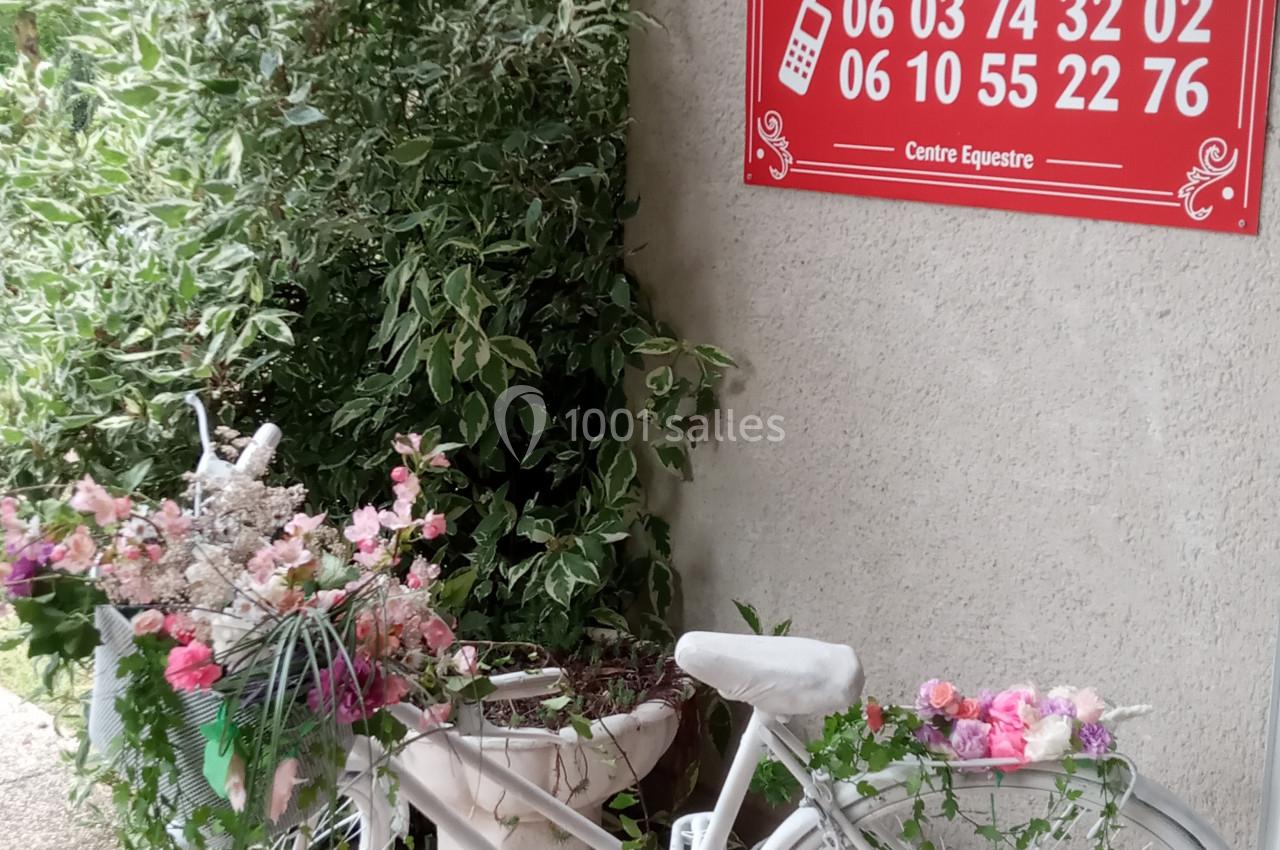 Vélo blanc décoré de fleurs devant un panneau rouge indiquant la location d'une salle de réception.