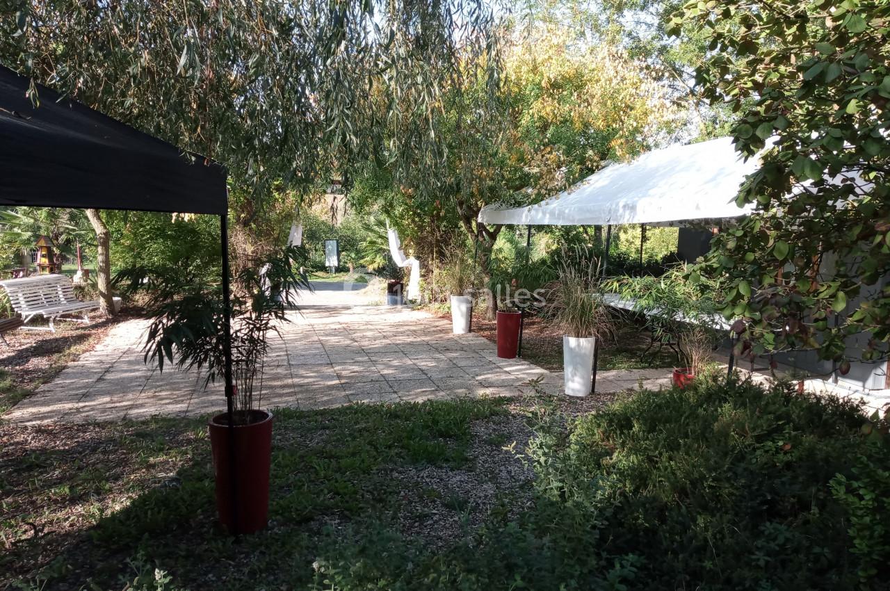 Allée pavée ombragée entourée de végétation, avec des tentes blanches et noires et des pots de plantes.