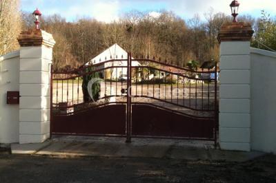 Portail en fer forgé rouge avec piliers blancs, donnant sur une allée menant à une maison entourée d'arbres.