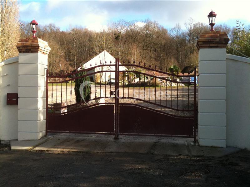 Portail en fer forgé rouge avec piliers blancs, donnant sur une allée menant à une maison entourée d'arbres.