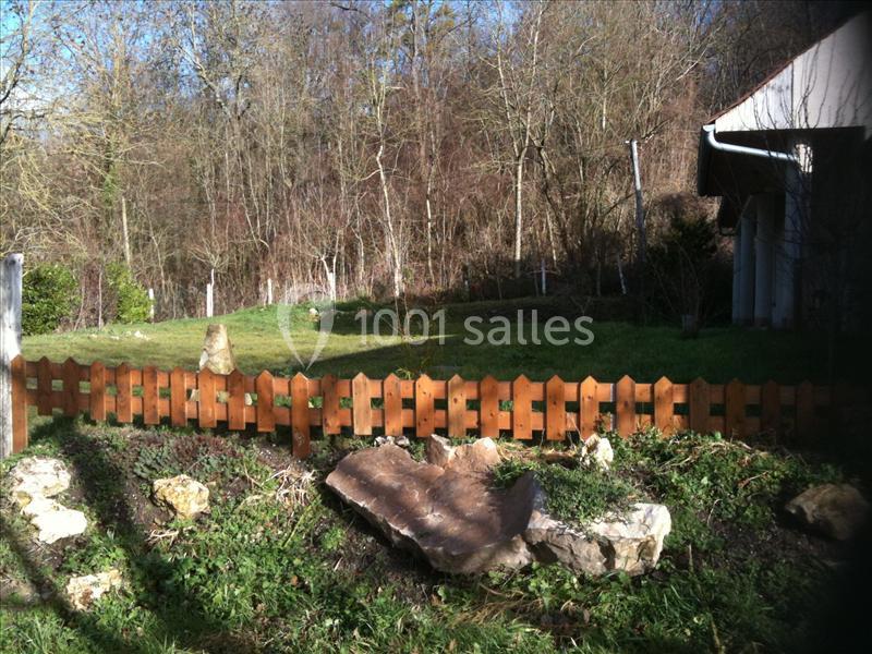 Clôture en bois devant un jardin avec des arbres en arrière-plan et une maison partiellement visible à droite.