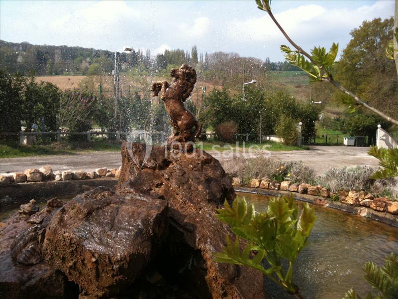 Statue de lion sur un rocher au centre d'une fontaine, entourée de végétation et d'un paysage rural.