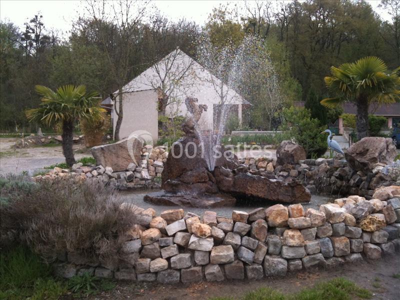 Fontaine décorative en pierre entourée d'un muret, avec jets d'eau, palmiers et végétation en arrière-plan.