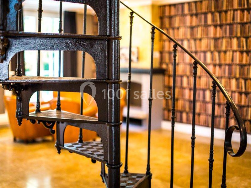 Escalier en colimaçon en métal noir dans un intérieur chaleureux avec mur décoré de faux livres et fauteuils en cuir.