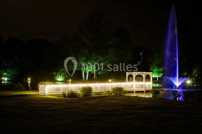Pont éclairé et kiosque en bois près d'une fontaine illuminée, entourés d'arbres dans un parc nocturne.