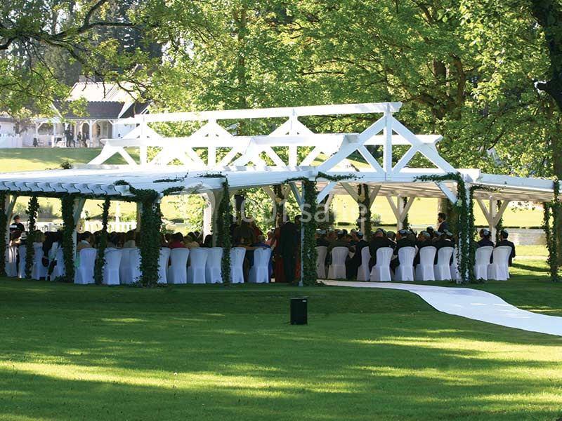 Structure blanche en plein air décorée de verdure, abritant des invités assis à des tables, dans un cadre verdoyant.