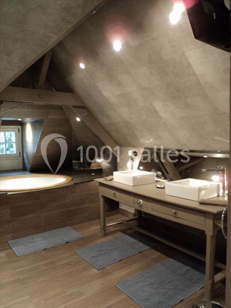 Salle de bain mansardée avec deux vasques sur un meuble en bois, baignoire encastrée et éclairage tamisé.