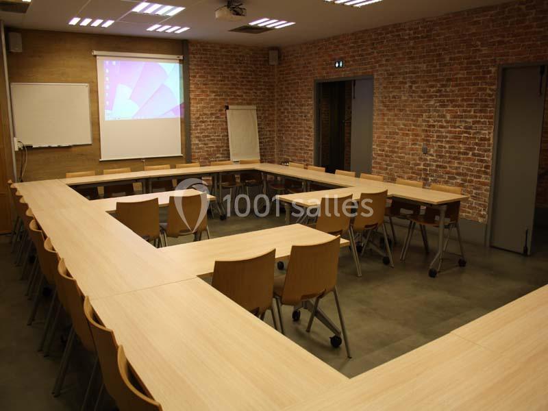 Salle de réunion avec tables disposées en U, chaises en bois, mur en briques et écran de projection allumé.