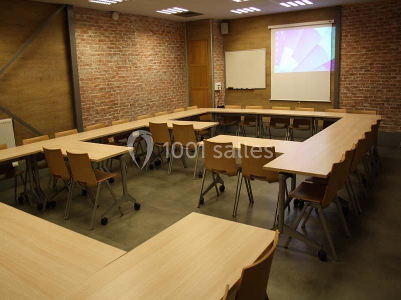 Salle de réunion avec tables disposées en U, chaises en bois, mur en briques et écran de projection allumé.