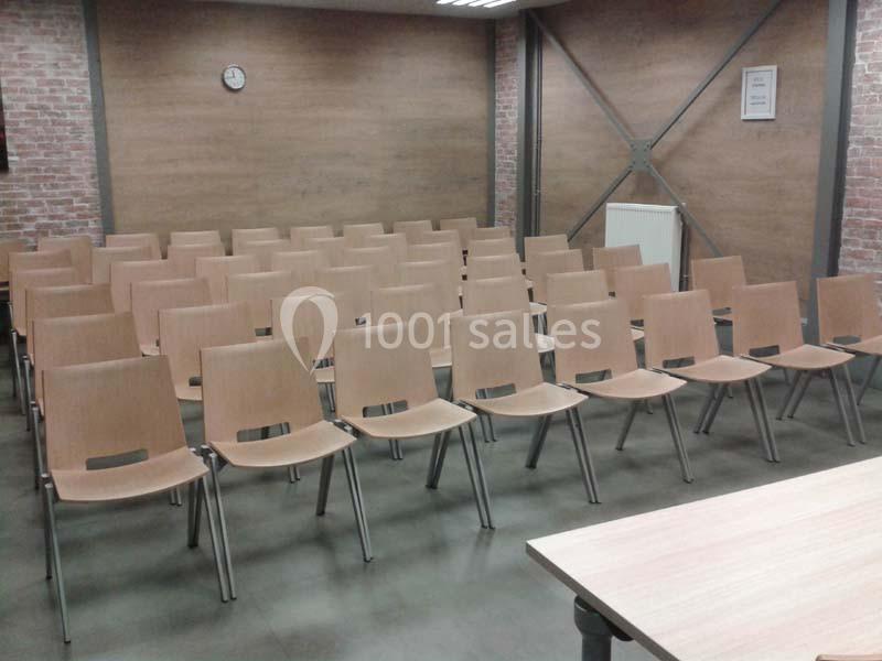 Salle de réunion avec plusieurs rangées de chaises en bois vides, mur en briques et horloge murale visible.