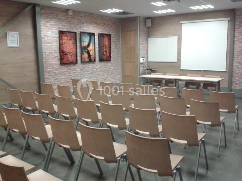 Salle de conférence vide avec des rangées de chaises en bois, un écran de projection et des tableaux accrochés au mur.