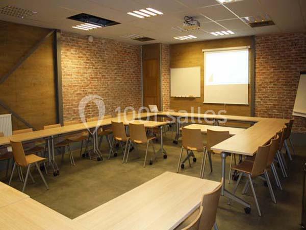 Salle de réunion vide avec tables en bois disposées en U, chaises, tableau blanc et mur en briques apparentes.