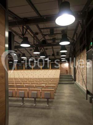 Salle de conférence vide avec des rangées de sièges en bois, éclairée par des lampes suspendues au plafond industriel.