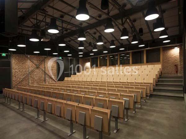 Salle de conférence vide avec des rangées de sièges en bois, murs en briques et plafond équipé de luminaires suspendus.