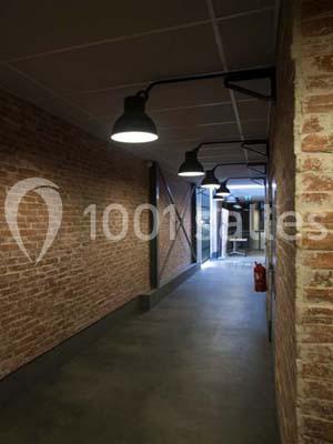 Couloir avec murs en briques, sol en béton, éclairage suspendu et porte métallique au fond.