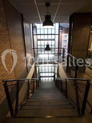 Escalier intérieur en métal avec rambardes, éclairé par une grande baie vitrée et des murs en briques apparentes.