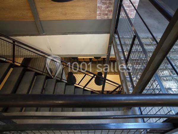Escalier en métal vu de haut, avec des murs en briques et bois, menant à un espace avec des tables et des chaises.