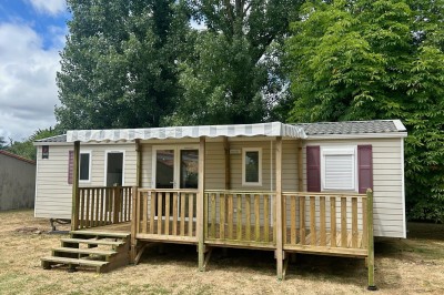 Mobil-home avec terrasse en bois, situé sur une pelouse, entouré d'arbres et partiellement ombragé.