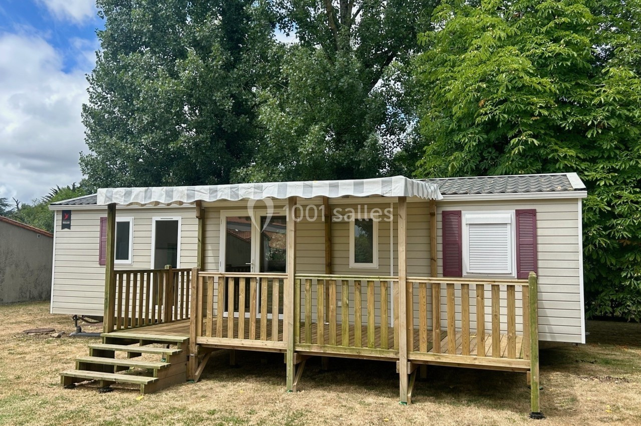 Mobil-home avec terrasse en bois, situé sur une pelouse, entouré d'arbres et partiellement ombragé.