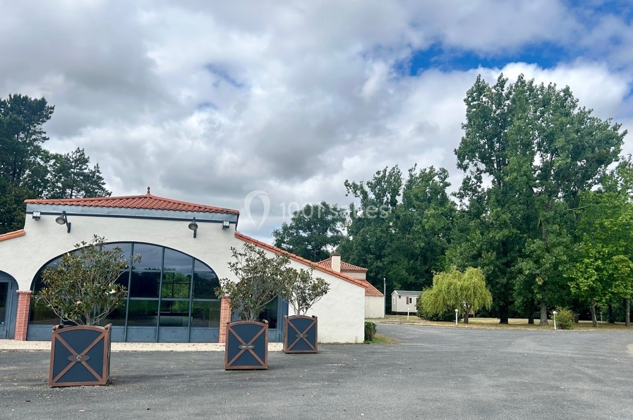 Bâtiment blanc avec toit en tuiles rouges, grande baie vitrée et arbres en arrière-plan sous un ciel nuageux.