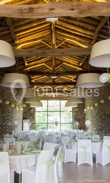Salle de réception avec murs en pierre, poutres apparentes, tables décorées et grandes fenêtres laissant entrer la lumière.