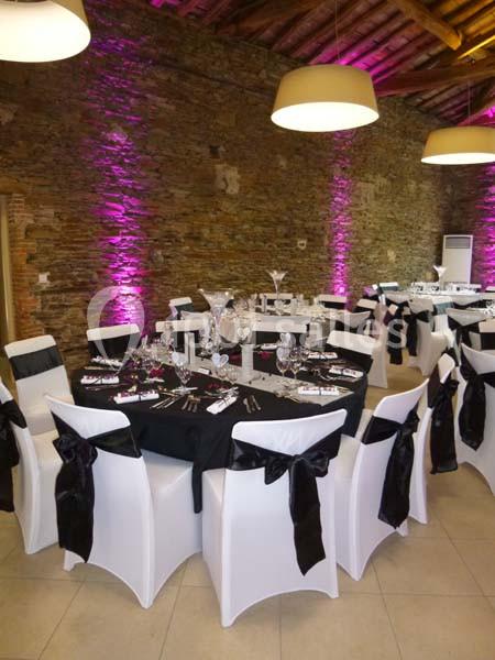 Salle de réception avec murs en pierre, tables rondes dressées, nappes noires et éclairage rose.