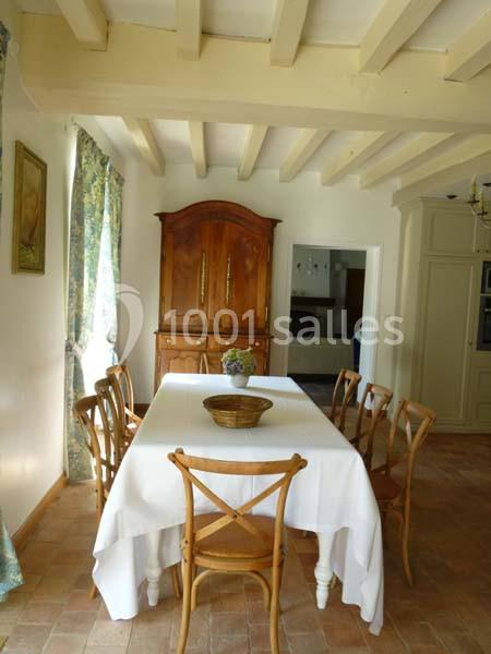 Salle à manger lumineuse avec table rectangulaire couverte d'une nappe blanche, chaises en bois et buffet en arrière-plan.