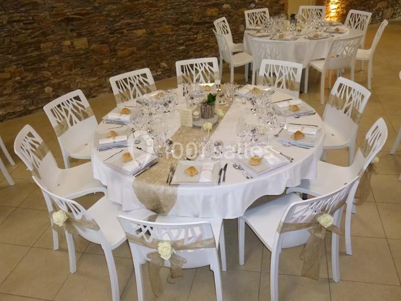 Table ronde décorée pour un événement, avec nappes blanches, chemins de table beiges, vaisselle et chaises ornées de rubans.