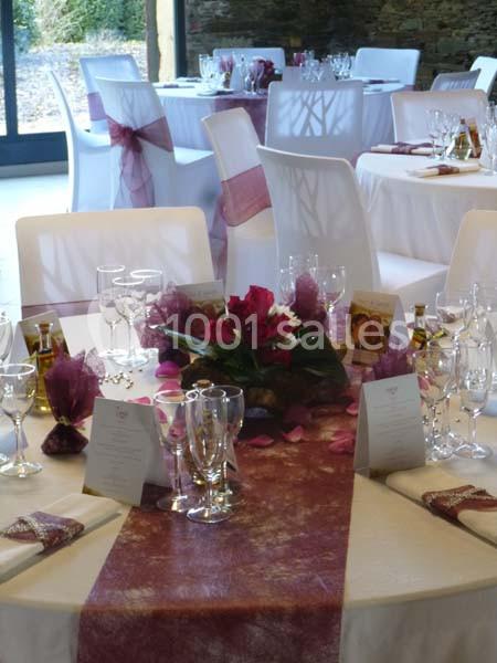 Salle de réception décorée avec des tables dressées, nappes blanches et chemins de table bordeaux, ornées de fleurs.