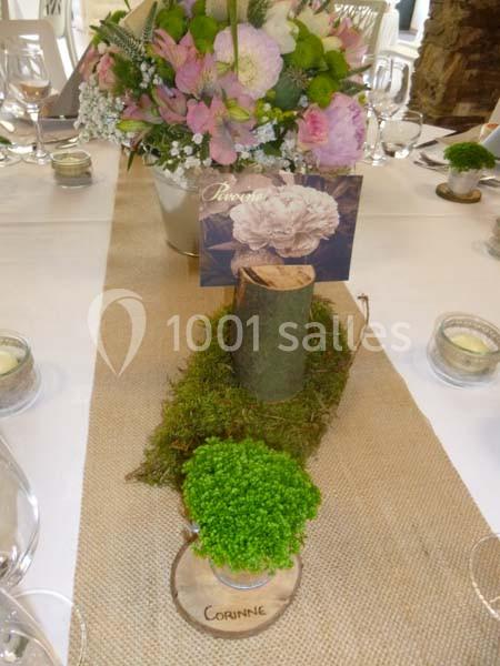 Centre de table avec fleurs roses, mousse, rondin de bois et marque-place en bois sur une nappe en toile de jute.
